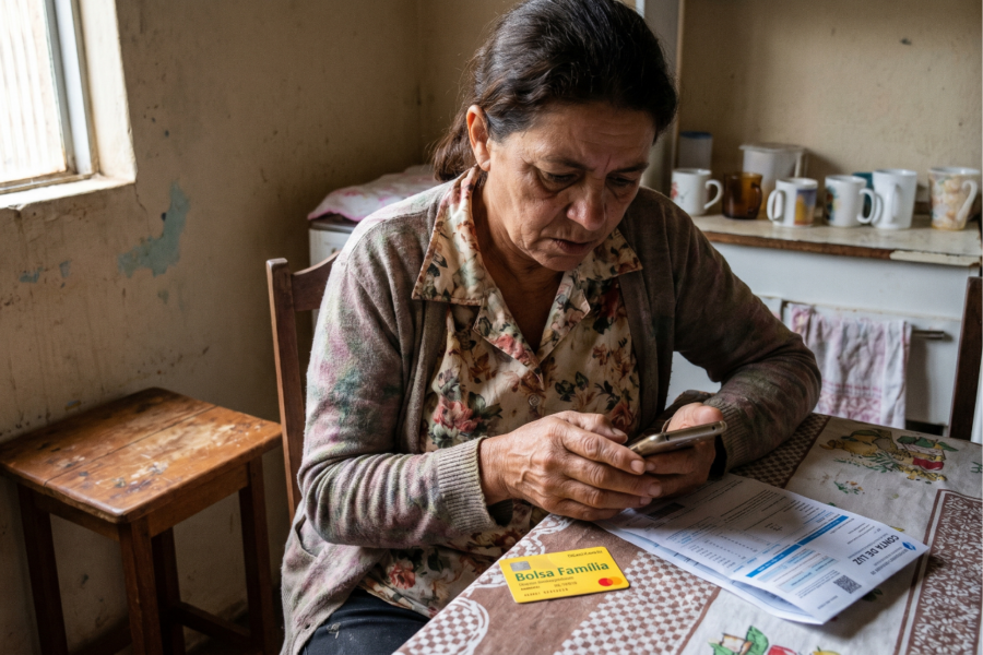 Mulher madura sentada sozinha à mesa da cozinha olhando preocupada para o celular, com um cartão do benefício e uma conta de luz ao lado.